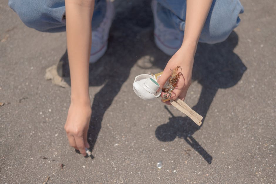 A person crouching on a paved outdoor surface, holding a small wooden stick with a partially broken white plastic object attached to it, which appears to be a discarded container or packaging. The individual's arms and hands are visible, with the left hand resting on the ground and the right hand gripping the object. The person is dressed in light blue jeans and grey shoes, and the scene is illuminated by natural light, casting shadows on the textured asphalt. The background is out of focus, emphasizing the person's hands and the rubbish they are holding, illustrating a scene of litter collection or waste management outdoors, relevant to private rubbish removal services such as those offered by Rubbish Removal Chiswick.