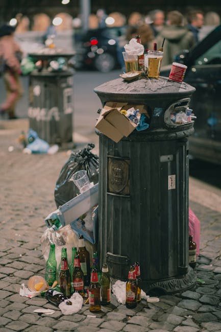 A black, cylindrical rubbish bin with a domed lid, situated on a cobblestone pavement in an outdoor urban setting. The bin's surface shows signs of wear with visible scratches and patches of rust. It is surrounded by scattered trash, including several empty glass bottles, crumpled paper, and discarded food packaging. On top of the lid, there are used paper cups with plastic lids, some containing remnants of drinks, alongside a crumpled paper napkin. A cardboard box is partially lodged into the side opening of the bin, with more litter spilling onto the ground. In the background, a black car is parked to the right, and a grey trash bag is leaning against the bin. There are blurred figures of people walking and another rubbish bin further away, indicating a busy street environment typical of city areas where independent waste disposal occurs. This scene highlights issues related to open trash and the need for effective rubbish removal services in outdoor public spaces, such as those offered by Rubbish Removal Chiswick.