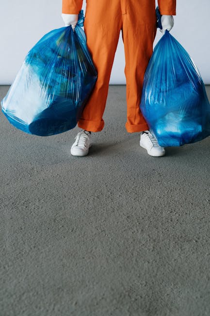 The image shows a person wearing orange overalls and white sneakers standing on a flat, grey concrete surface. The individual is holding two large, semi-transparent blue plastic rubbish bags, one in each hand, which are filled with waste materials that are faintly visible through the plastic. The background is plain and light-colored, with no identifiable objects or environmental features, suggesting an indoor or neutral setting. The scene emphasizes the act of waste collection or disposal, aligning with private rubbish removal services such as those provided by Rubbish Removal Chiswick. The bags appear to be sturdy and suitable for carrying household or light commercial rubbish, while the person's stance indicates they are in the process of transporting waste for disposal or collection. The overall composition underlines the themes of waste management, independent collection, and on-site clearance, typical in non-reliance on local authority rubbish services.