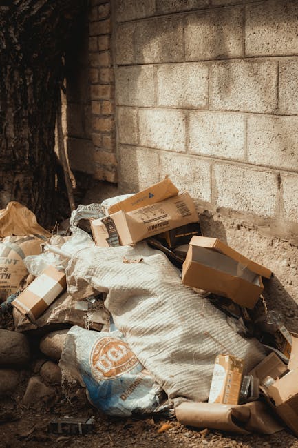 The image shows a person wearing orange overalls and white sneakers standing on a flat, grey concrete surface. The individual is holding two large, semi-transparent blue plastic rubbish bags, one in each hand, which are filled with waste materials that are faintly visible through the plastic. The background is plain and light-colored, with no identifiable objects or environmental features, suggesting an indoor or neutral setting. The scene emphasizes the act of waste collection or disposal, aligning with private rubbish removal services such as those provided by Rubbish Removal Chiswick. The bags appear to be sturdy and suitable for carrying household or light commercial rubbish, while the person's stance indicates they are in the process of transporting waste for disposal or collection. The overall composition underlines the themes of waste management, independent collection, and on-site clearance, typical in non-reliance on local authority rubbish services.