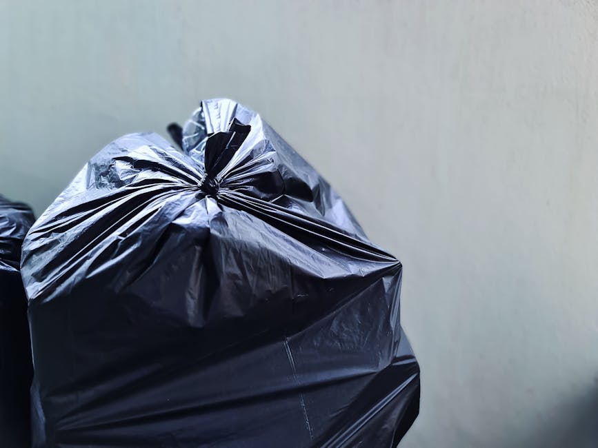 Close-up image of a large black plastic rubbish bag tightly sealed at the top with creases and folds visible on its surface, positioned in the foreground against a neutral, light-colored plain wall background. The bag appears to be made of high-density polyethylene, with a slightly reflective, crinkled texture that suggests it is filled with waste materials. Surrounding the main bag, part of another similar black bag is partially visible, stacked or placed nearby, indicating a collection of waste ready for disposal. The scene is well-lit with soft, natural or artificial lighting, without shadows, emphasizing the bag's glossy finish and detailed surface. The overall environment suggests an indoor or sheltered outdoor setting where private rubbish collection or waste removal services, such as those offered by Rubbish Removal Chiswick, might be carried out as part of on-site or alternative waste handling procedures, set in a context of preparing waste for collection outside a residential or commercial property.