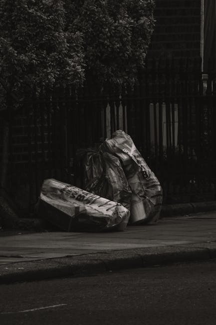 Two large black plastic garbage bags filled with waste are positioned on the pavement near a black metal fence. The bags appear to contain household rubbish and are slightly crumpled with visible creases on their surfaces. One bag is leaning against the other, with the top of the bags gathered and tied at the open ends. The scene is set outdoors on a street, with a dark, leafy hedge or tree in the background, suggesting an urban or residential area. The surrounding environment includes a section of paved road and a curb, with natural light illuminating the scene, highlighting the textures and folds of the bags. The image depicts an informal rubbish collection, possibly representing private waste removal in a suburban setting, which aligns with the services offered by Rubbish Removal Chiswick for alternative waste handling solutions.