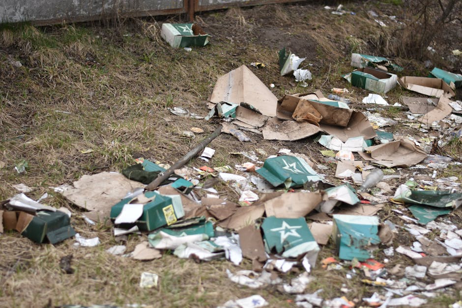 A collection of discarded cardboard boxes, many crushed or torn, scattered across a grassy area near a fence. The boxes are primarily green and brown, with visible logos and labels indicating they previously contained household goods or packaging materials. Some boxes are partially flattened, while others are still upright or leaning. The ground is uneven, with patches of soil and sparse grass, and there are small pieces of paper and plastic debris mixed in among the boxes. In the background, a wooden fence runs horizontally, and there are some dried, leafless bushes and twigs along the fence line. The scene appears to be outdoors, possibly in a suburban or urban setting, illustrating improper waste disposal or illegal dumping, which a professional waste removal service like Rubbish Removal Chiswick might assist with by providing controlled, environmentally responsible rubbish collection. Natural lighting suggests daytime, with no visible weather effects.