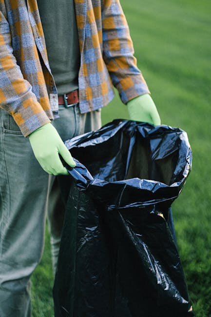 Two large black plastic garbage bags filled with waste are positioned on the pavement near a black metal fence. The bags appear to contain household rubbish and are slightly crumpled with visible creases on their surfaces. One bag is leaning against the other, with the top of the bags gathered and tied at the open ends. The scene is set outdoors on a street, with a dark, leafy hedge or tree in the background, suggesting an urban or residential area. The surrounding environment includes a section of paved road and a curb, with natural light illuminating the scene, highlighting the textures and folds of the bags. The image depicts an informal rubbish collection, possibly representing private waste removal in a suburban setting, which aligns with the services offered by Rubbish Removal Chiswick for alternative waste handling solutions.